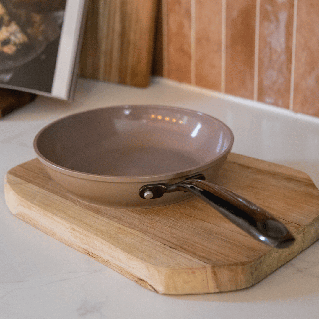 Taupe frying pan with black handle on wooden cutting board in kitchen setting.