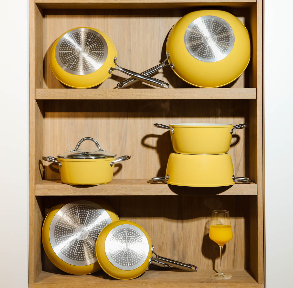 9-piece yellow cookware set with stainless steel bottoms and glass lids, displayed in a wooden cabinet.