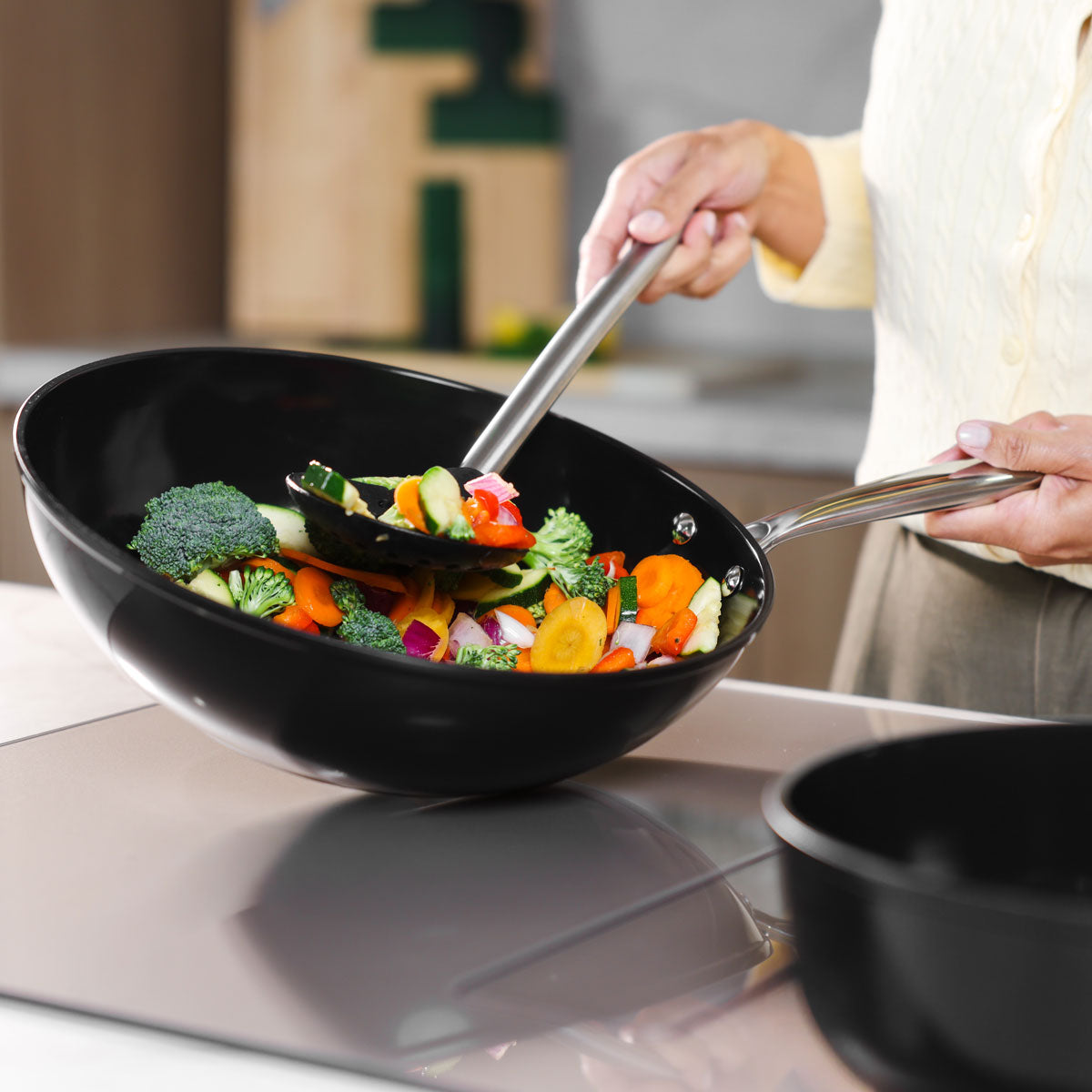 Black wok pan 30 cm with stainless steel handle, stir-fry vegetables inside, induction cooktop.