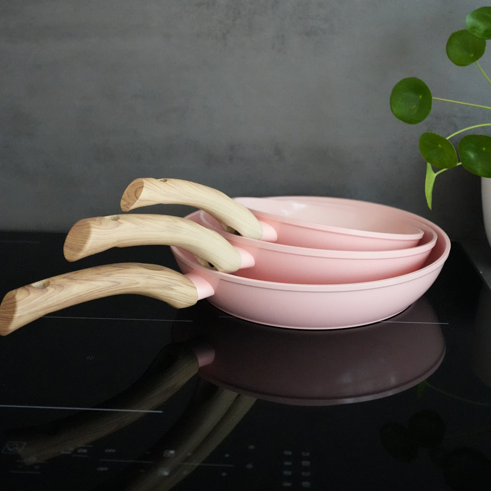 Set van drie roze koekenpannen met houten handvatten, gestapeld op een zwarte inductiekookplaat.