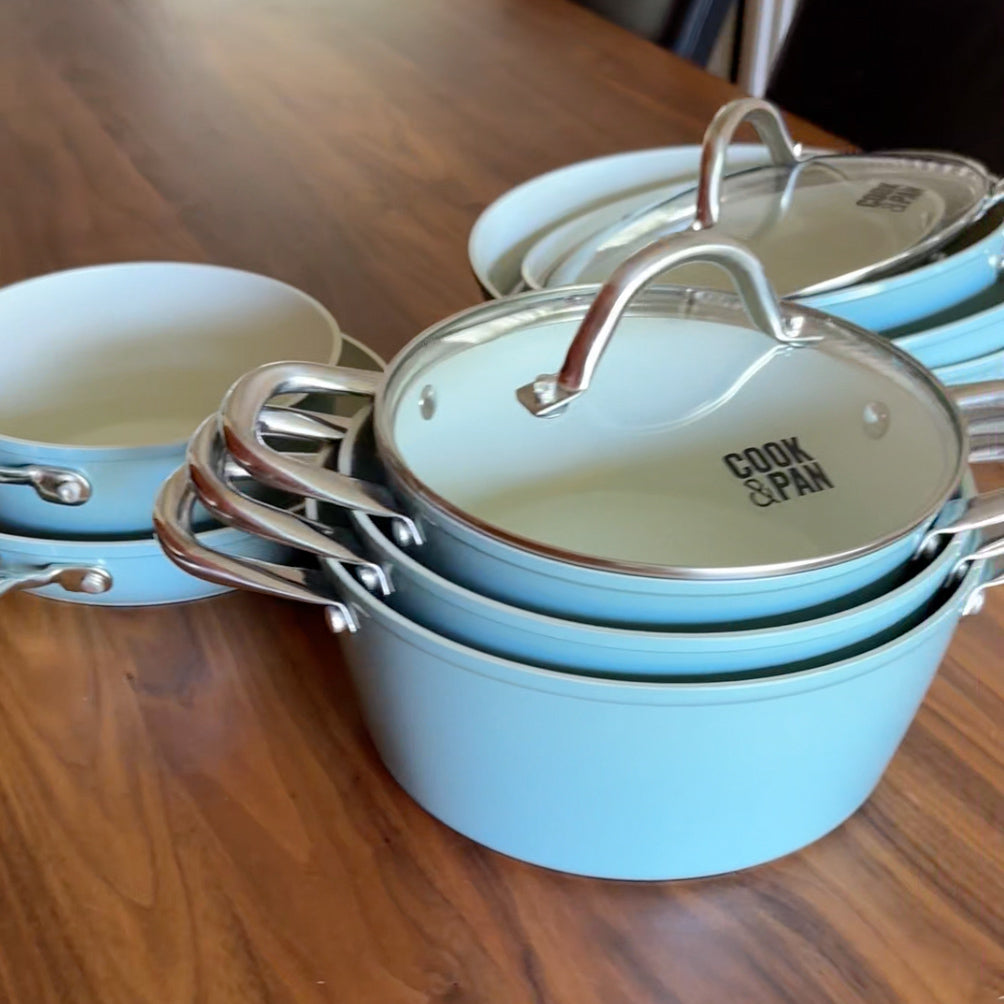Batterie de cuisine bleu clair neuf pièces avec couvercles en verre et poignées en acier inoxydable, empilée sur une table en bois.