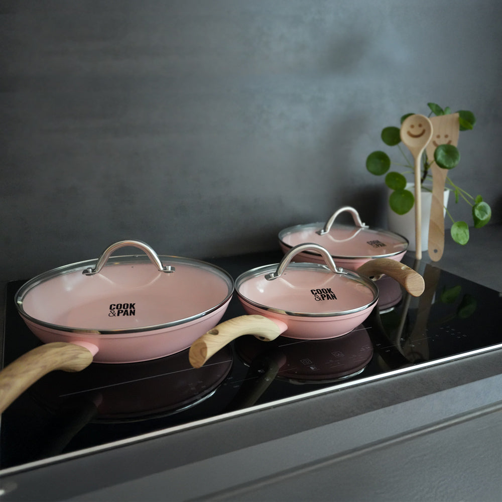 Set de poêles à frire rose de 3 pièces avec couvercles en verre et poignées en bois clair sur plaque de cuisson noire.