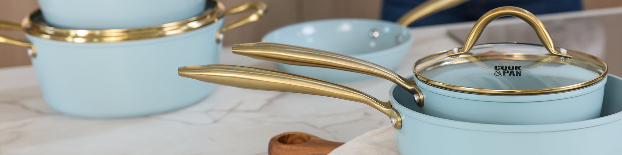 Lichtblauwe Cook & Pan pannen met gouden handvatten en glazen deksels op een marmeren aanrecht