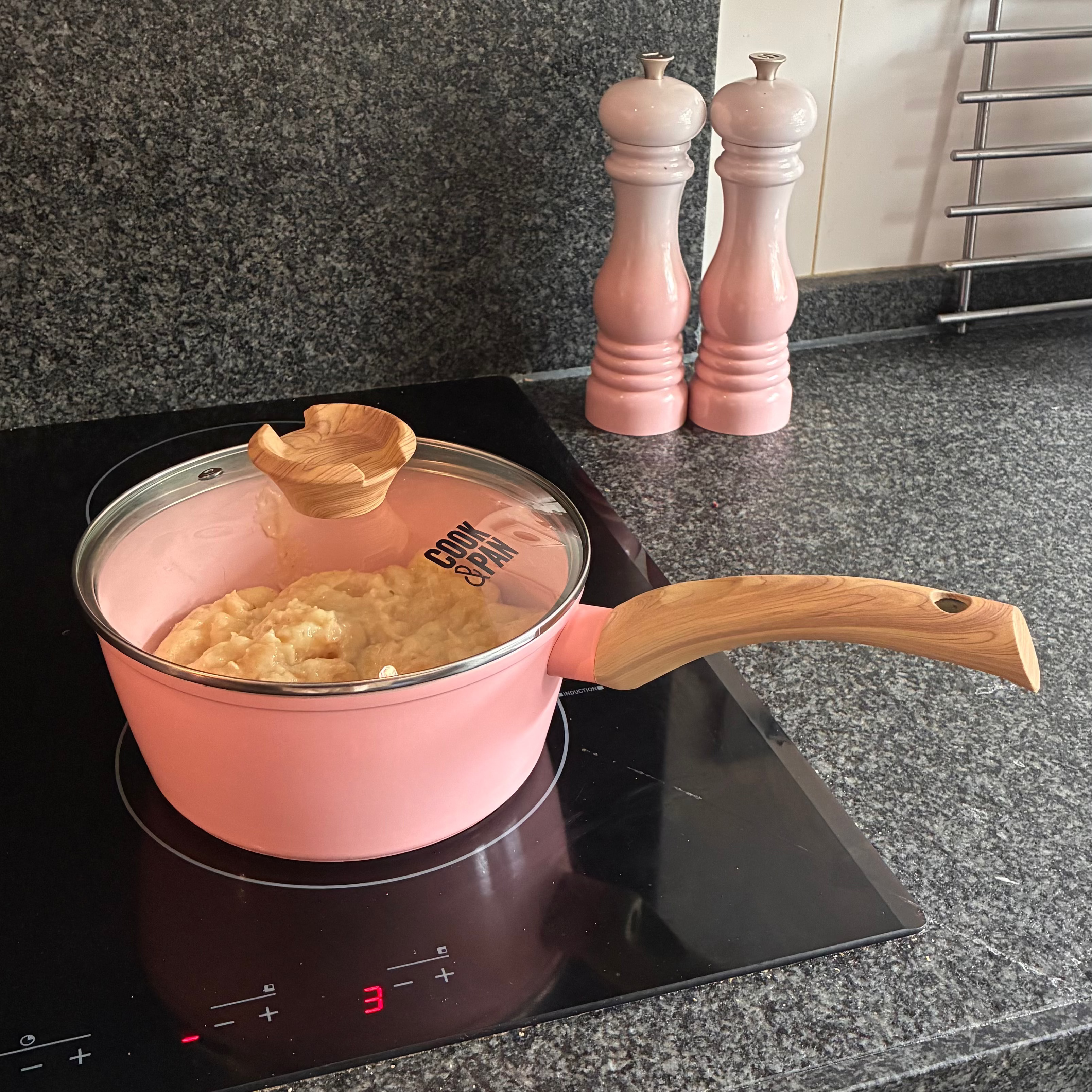 Batterie de cuisine rose avec couvercle en verre et poignée en bois, casserole remplie de flocons d’avoine sur plaque de cuisson.