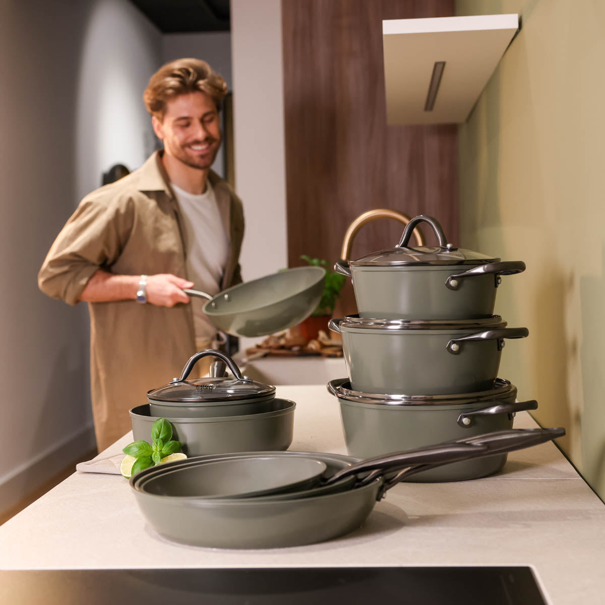 Dark green 20 cm frying pan with black handle lies in front of stack of pots and pans with glass lids.