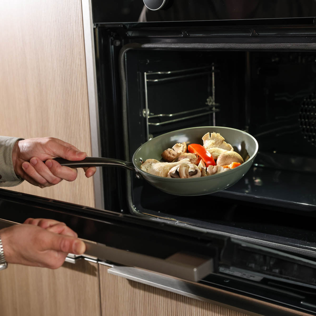 Dunkelgrüne Pfanne mit Antihaftbeschichtung und schwarzem Griff, gefüllt mit Gemüse und Hähnchen, im schwarzen Ofen platziert.