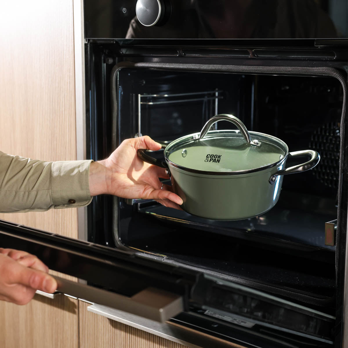 Batterie de cuisine vert foncé avec poignées en acier inoxydable et couvercle en verre placée dans un four.