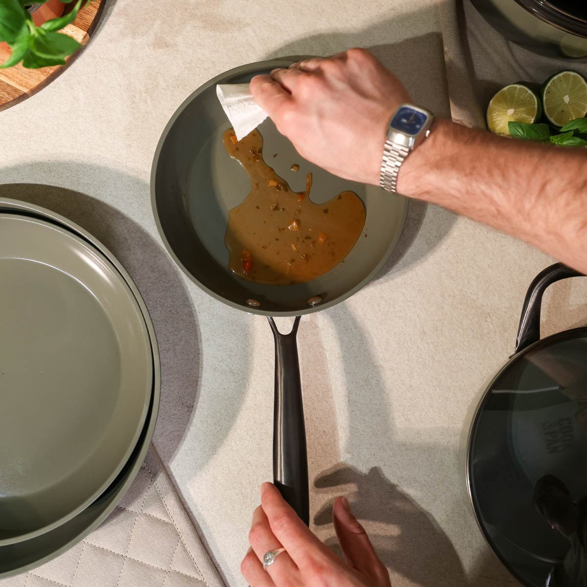 Poêle vert foncé avec revêtement antiadhésif dans laquelle une sauce est versée, à côté d'une pile de casseroles et poêles et d'un couvercle.