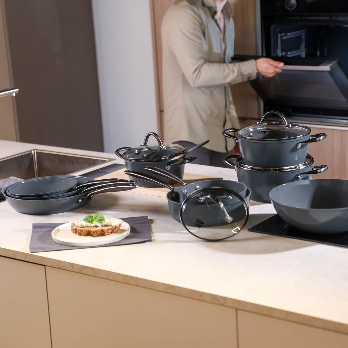 Set of black frying pans with glass lids on kitchen counter, with man opening oven.