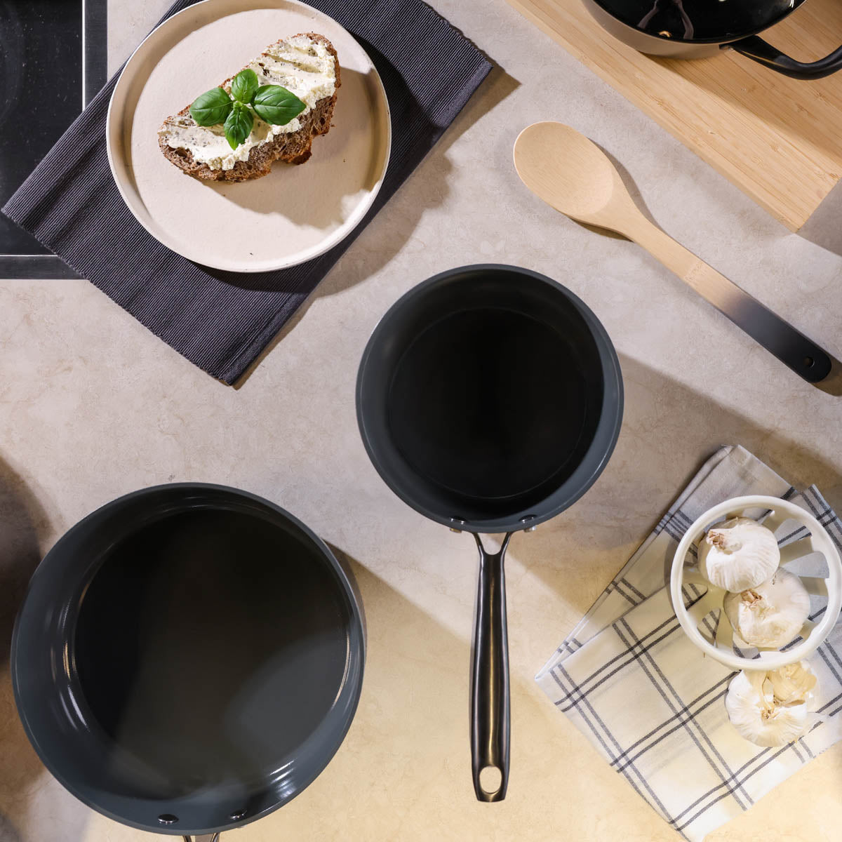 Black non-stick frying pan and saucepan with wooden spoon on marble surface.