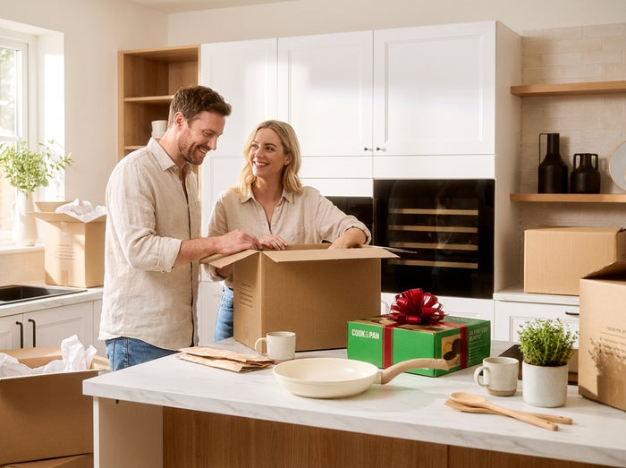 Un couple déballe des cartons dans une nouvelle cuisine, avec une poêle crème et une boîte Cook & Pan verte à nœud rouge sur le plan de travail
