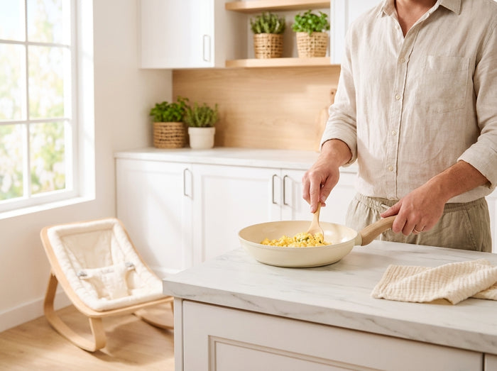 Un parent cuit un œuf dans une poêle crème dans une cuisine claire, un transat bébé en arrière-plan