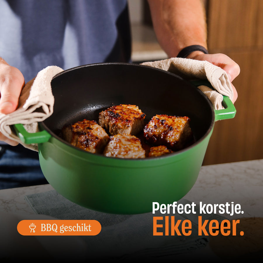 Cocotte en fonte Go Green avec surface émaillée verte contenant des morceaux de viande grillée, tenue par deux mains avec un torchon en lin.