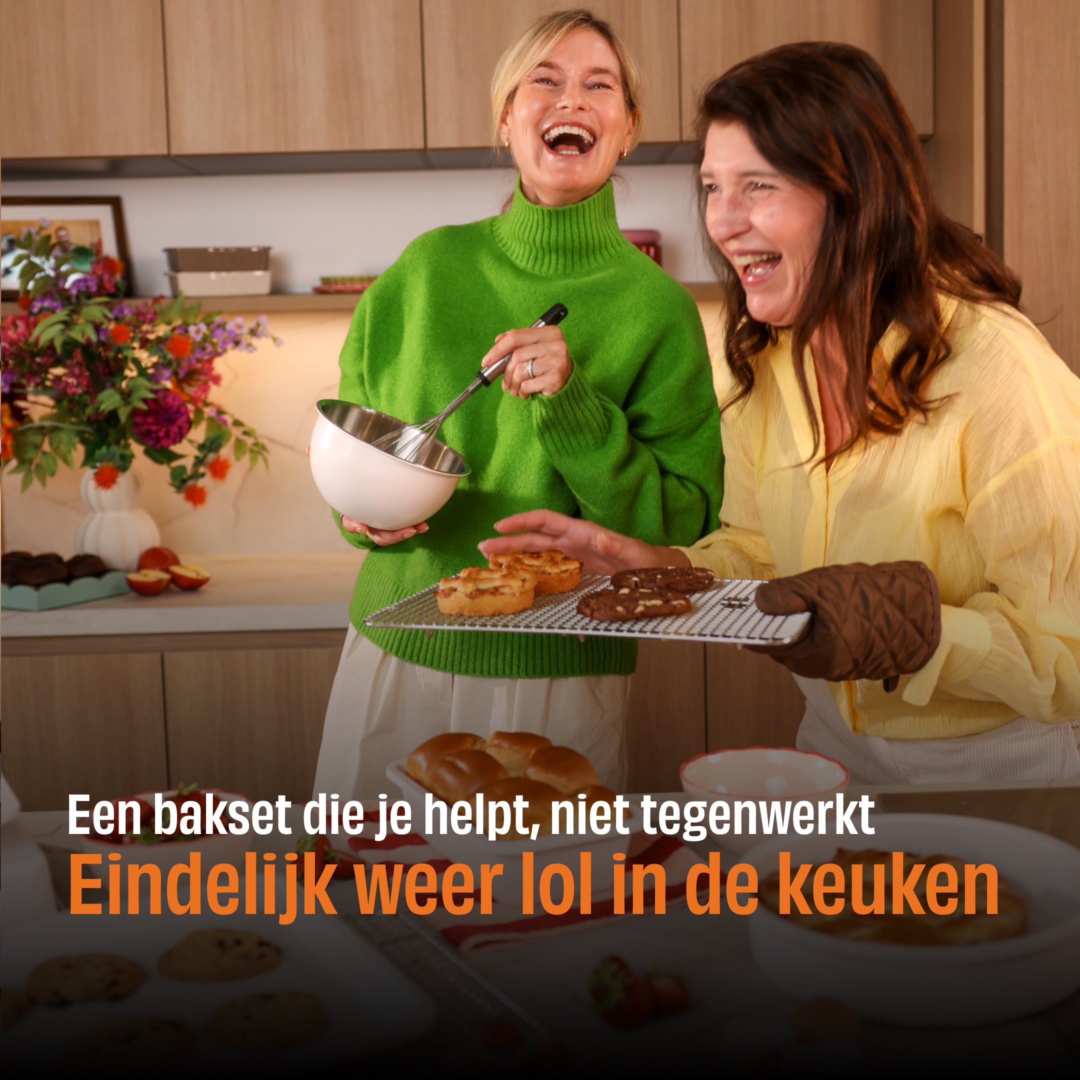 Twee vrouwen lachen in een keuken met Go Ivory bakset, waarvan een witte bakschaal met metalen handvat met gebak gevuld